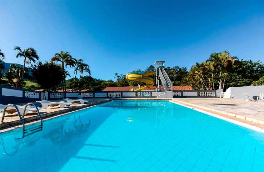 Durante o dia, piscina externa com palmeiras ao lado, tobogã amarelo, vegetação ao redor e céu azul