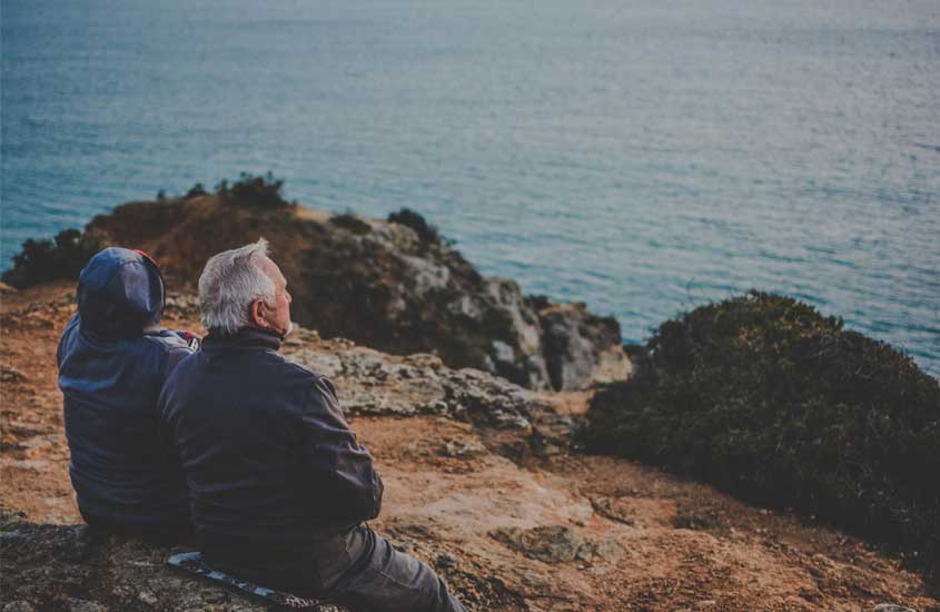 Durante o dia, dois idosos sentados em cima de uma pedra em frente o mar e a vegetação