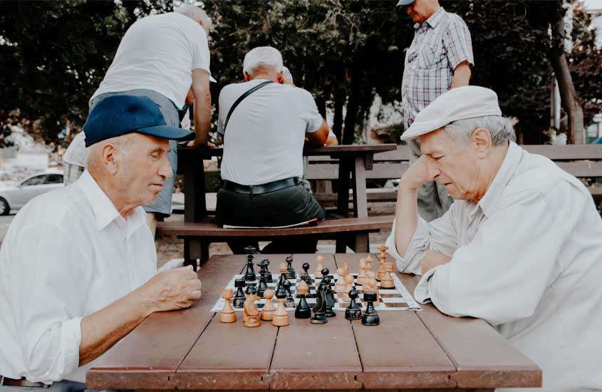 dois idosos jogando xadrez em mesa de praça com mais 3 homens e árvores ao fundo