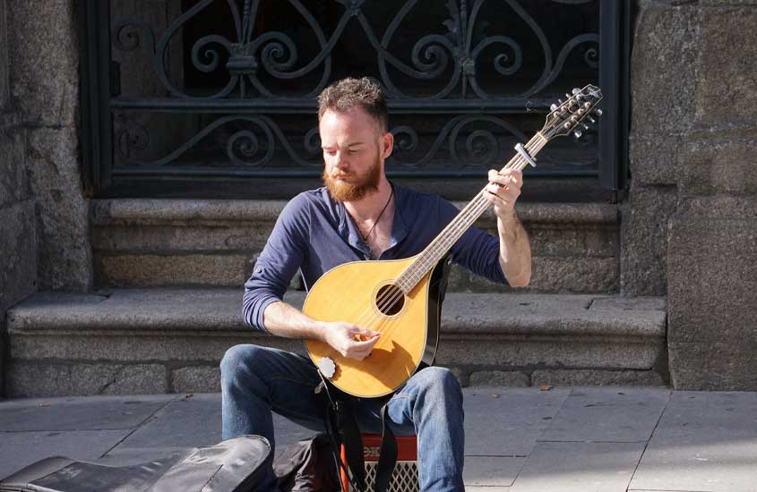 Em dia de sol, homem sentado na calçada tocando fado com guitarra portuguesa, ritmo que, de acordo com curiosidades sobre Portugal, é patrimônio da unesco