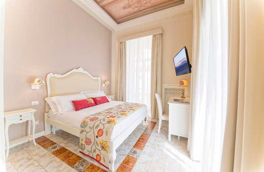 Quarto de hotel em Nápoles m tons neutros com detalhes laranja e rosa, pintura no teto, cama de casal, área de trabalho e televisão