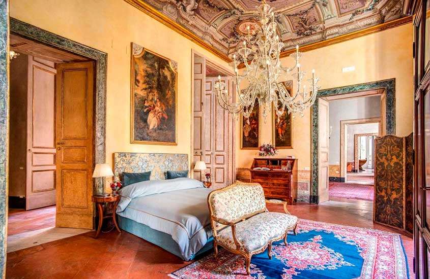 quarto luxuoso de um dos hotéis em Nápoles, todo decorado com obras de arte, pinturas no teto, tapete, lustre grande, cama de casal e cômoda