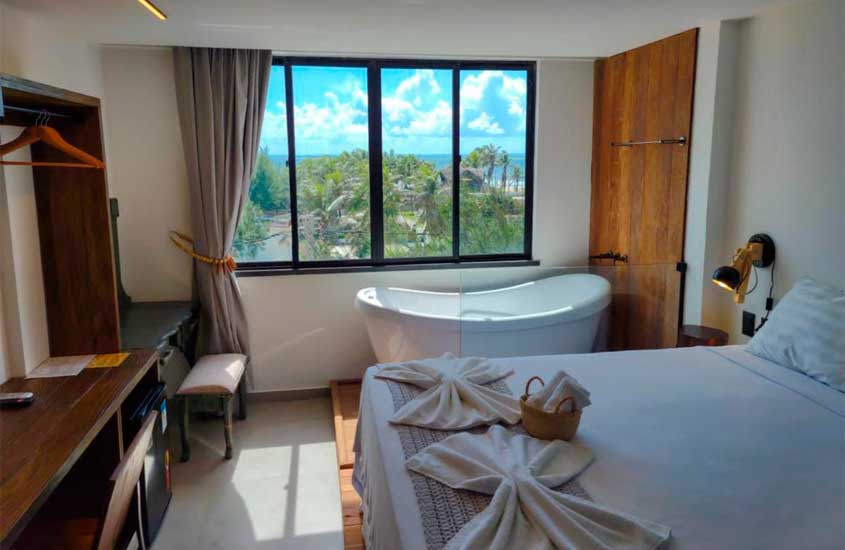 Em dia de sol, quarto de hotel em fortaleza com cama de casal, banheira, área de trabalho, luminária e janela grande com vista para o mar