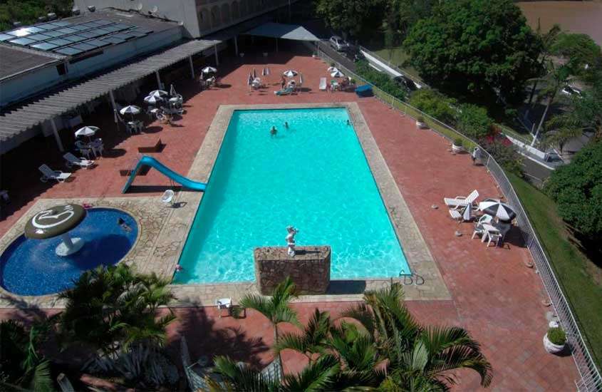 Durante o dia, vista aérea de área de piscina de hotel em aguas de lindoia com espreguiçadeiras brancas, escorregador azul, cascata pequena e vegetação ao redor