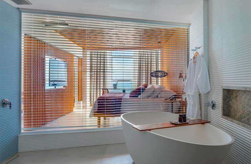 Em dia de sol, banheiro de suíte de hotel com banheira oval, chuveiro e roupões. Ao fundo, cama de casal, televisão, lustre e janela grande com vista para o mar