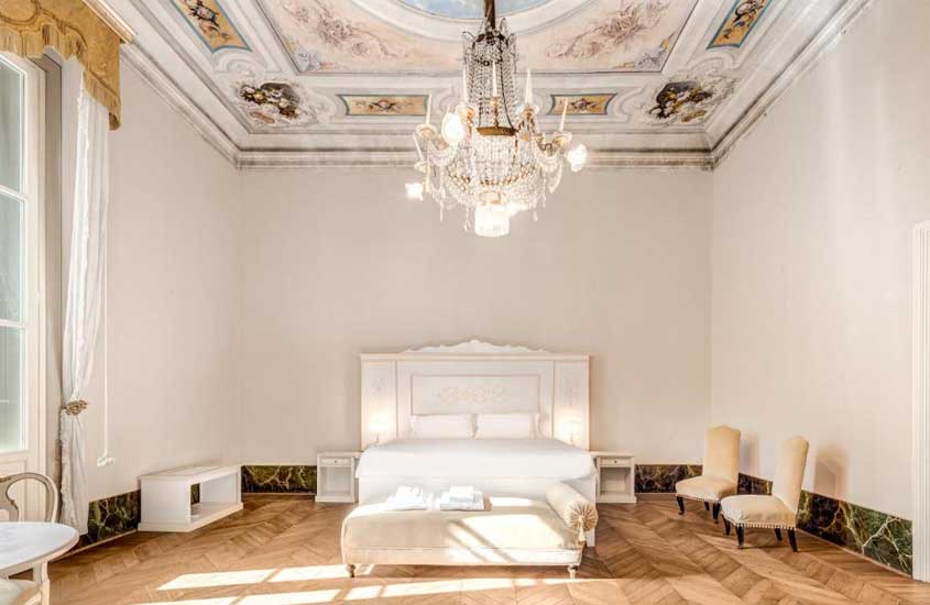 Quarto de hotel em Florença luxuoso, mobiliado com lustre grande, cadeiras e móveis brancos, chão de madeira e pintura no teto