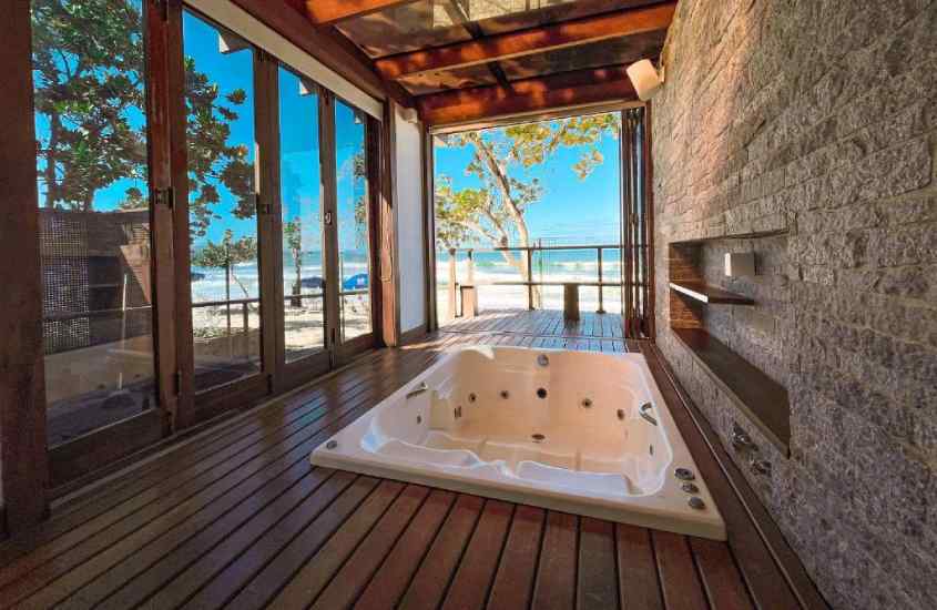 Área de banheira com deck de madeira, janelas grandes e vista da praia