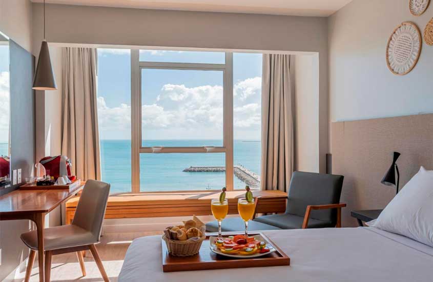 Em dia de sol com nuvens, tábua de café da manhã servida em cima de cama de casal de suíte de hotel com grandes janelas e vista para o mar