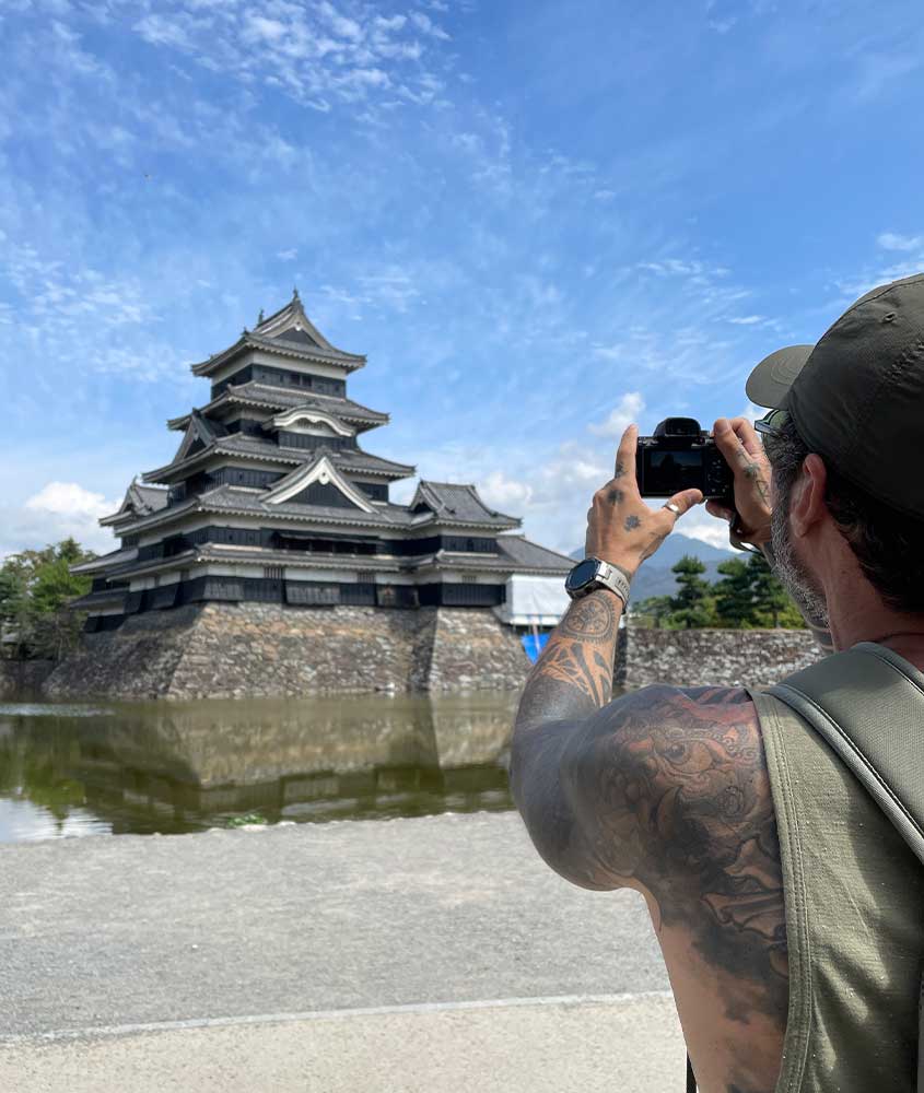 Durante um dia de sol, Vagner Alcantelado tirando foto do Castelo de Matsumoto com lago na frente e árvores ao redor
