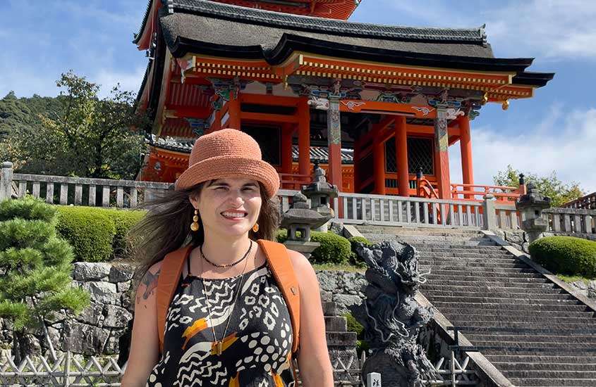 Em um dia de sol, Bárbara Rocha Alcantelado, criadora do blog melhores momentos da vida. em frente a um templo de Kyoto com plantas ao redor