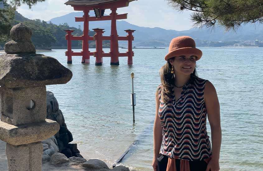 Em um dia de sol, Bárbara Rocha Alcantelado na Ilha de Miyajima com árvores ao redor e rio com escultura de madeira atrás