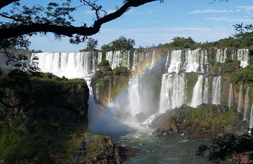 Em dia de céu azul, vista panorâmica de arco-íris entre as as cataratas do Iguaçu com árvores.