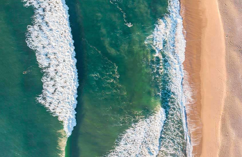 Vista aérea em dia ensolarado, mostrando o mar de verde azulado e as ondas arrebentando na faixa de areia seca e molhada, em Florianópolis.