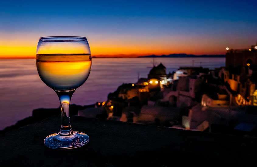 Durante pôr do sol, taça de vinho em cima de muro com vista para a cidade e o mar de Santorini, um dos melhores passeios na grecia