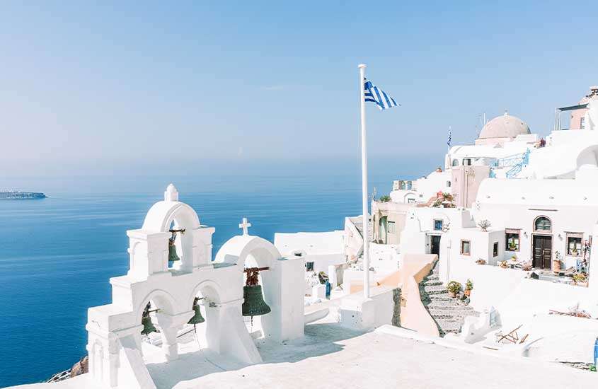 em dia ensolarado, bandeira da Grécia hasteada em cidade com diversas construções brancas e vista para o mar