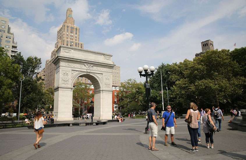em dia de sol, pessoas caminhando em praça de Nova York com réplica de arco do triunfo e árvores