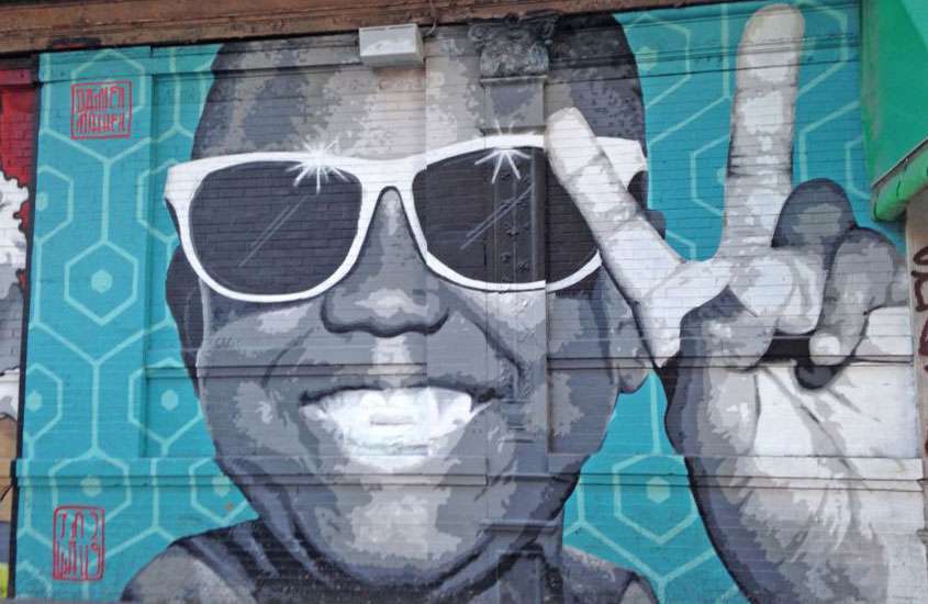 Durante o dia, parede pintada em tons de preto, branco, cinza e azul, com um homem negro sorridente e de óculos, fazendo o gesto da letra V com o dedo indicador e o dedo médio, simbolizando uma mensagem positiva ou vitória.