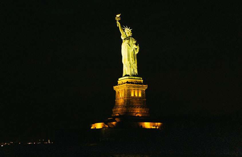 durante a noite estátua da liberdade iluminada com luz amarela, vista de cruzeiro, um dos melhores passeios em nova york