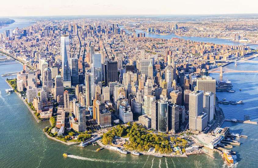 durante um dia ensolarado, vista aérea da cidade de nova york com edifícios, árvores, barcos e mar ao redor, vista de tour de helicópero, um dos melhores passeios em nova york
