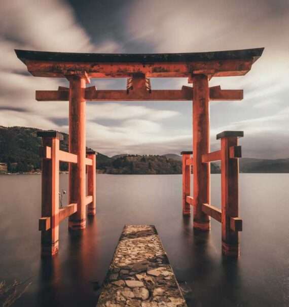 Durante um dia nublado, estrutura de madeira tradicional do Japão em cima de lago. Ao fundo, diversas montanhas
