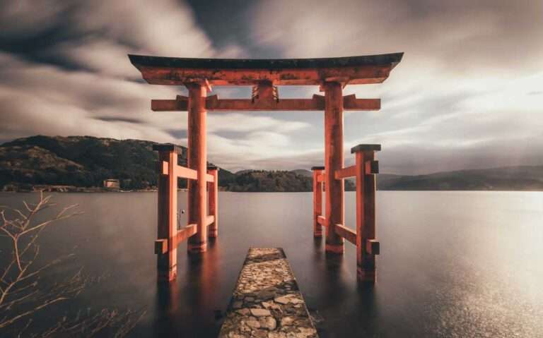 Durante um dia nublado, estrutura de madeira tradicional do Japão em cima de lago. Ao fundo, diversas montanhas