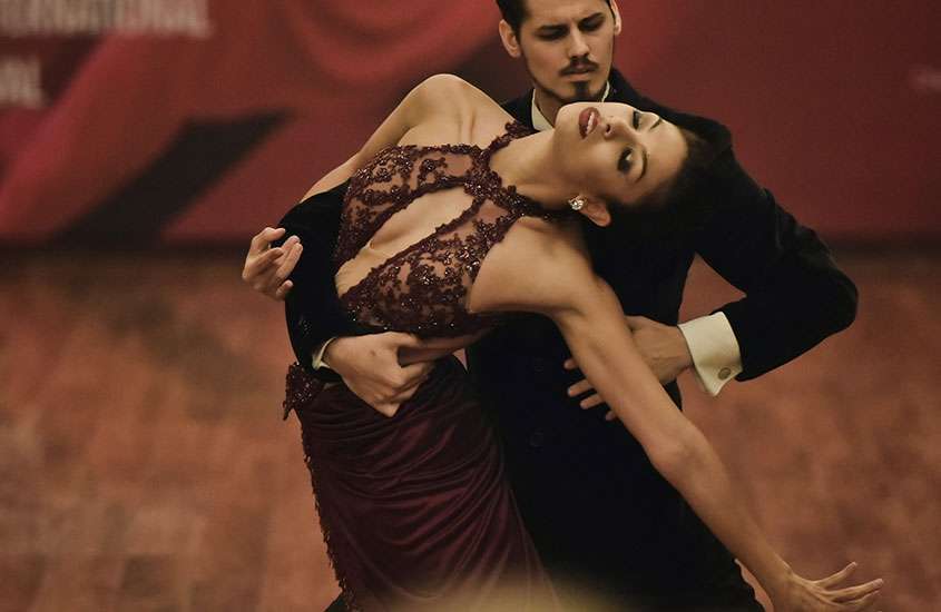 Casal dançando tango em um restaurante, a mulher vestindo um elegante vestido vinho e o homem usando um terno branco e preto