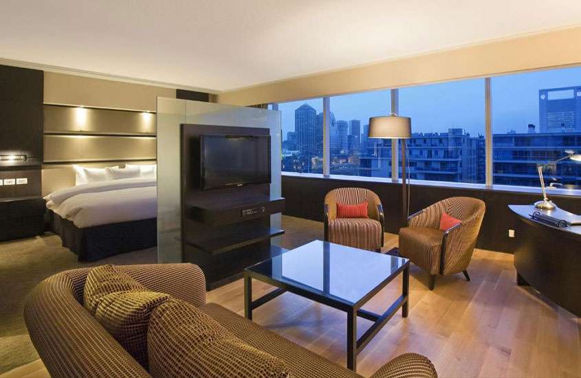 Durante o dia, quarto de hotel mobiliado com sofá, poltronas, tv e cama de casal e grandes janelas com vista da cidade