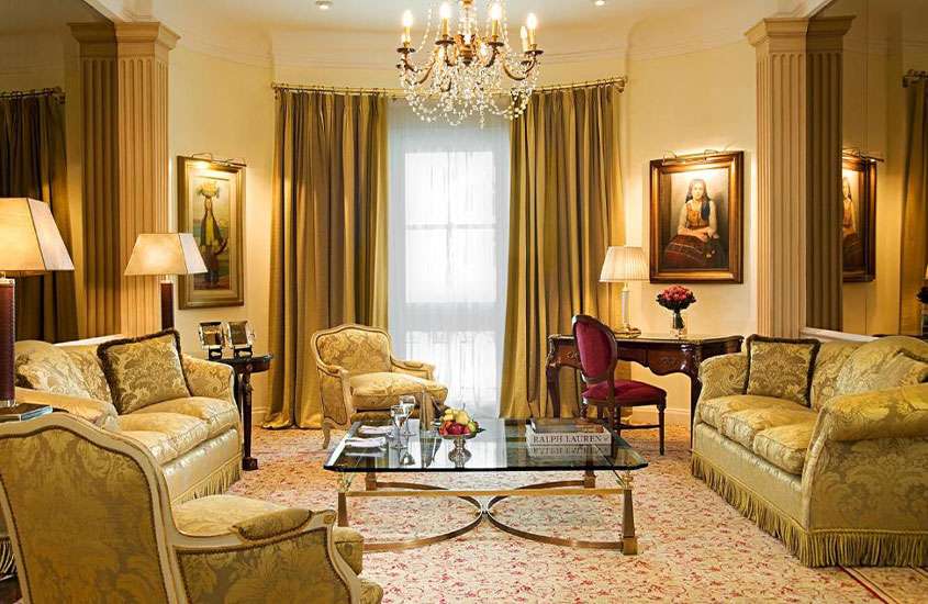 Sala de hotel com poltronas, sofás, mesa de madeira, obras de arte, lustre grande e janela com cortinas