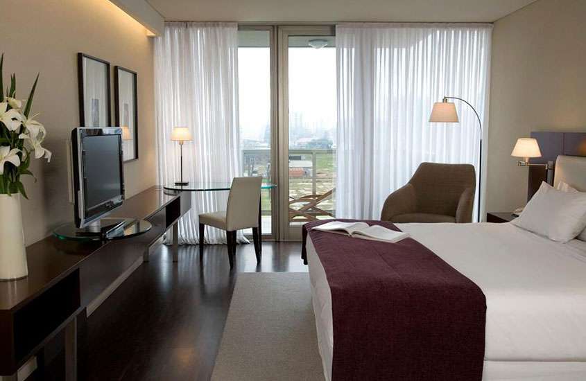 Vista de um quarto de hotel durante o dia, elegantemente mobiliado em tons neutros e escuros, apresentando uma cama de casal, uma poltrona confortável e grandes janelas com vista da cidade