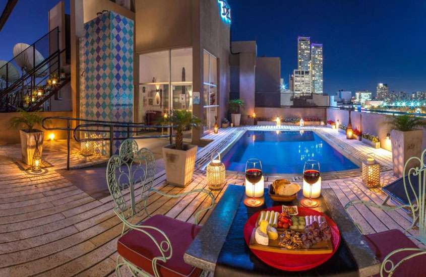Durante a noite, piscina e mesa posta com cadeiras de metal ao redor, em cobertura de hotel com vista da cidade
