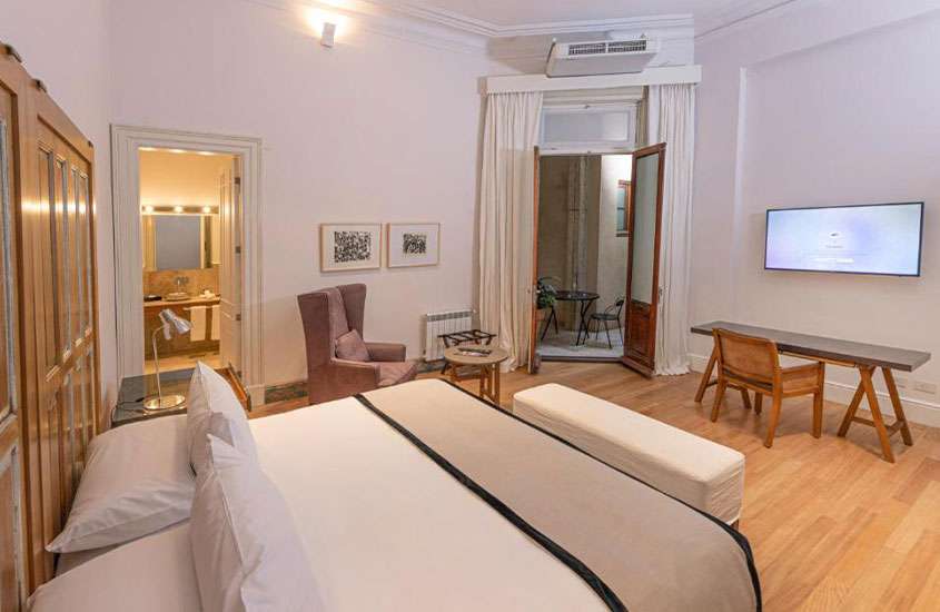 Quarto de hotel mobiliado em tons neutros e piso de madeira com cama de casal, armário, poltrona, ar-condicionado e mesa de trabalho