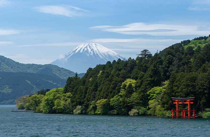 Visão panorâmica de árvores e montanhas ao redor de lago. Ao fundo, visão panorâmica do monte Fuji, um lugar para incluir no roteiro japão