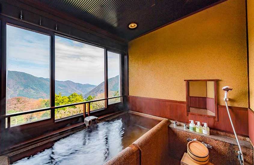 Durante o dia, área interna de hotel com banheira, produtos de corpo, secador e janela grande com vista das montanhas e das árvores de Hakone