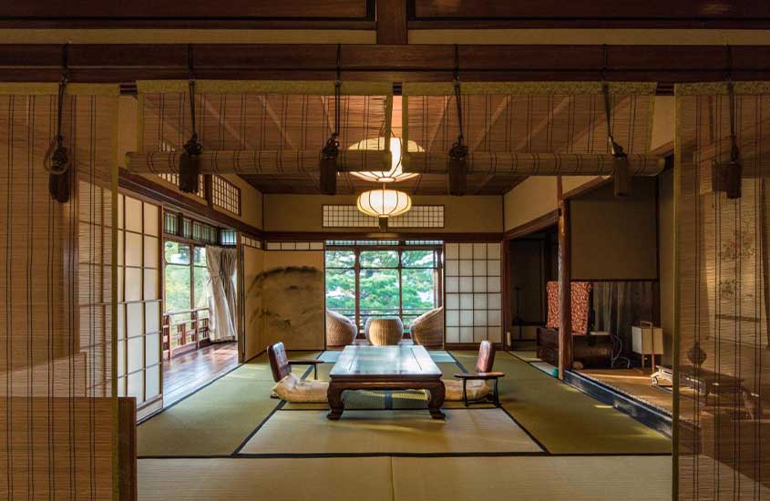 Vista interna de um hotel em Kyoto com mesas, cadeiras, poltronas, luminárias, lustres, televisão e grandes janelas com vista para a natureza