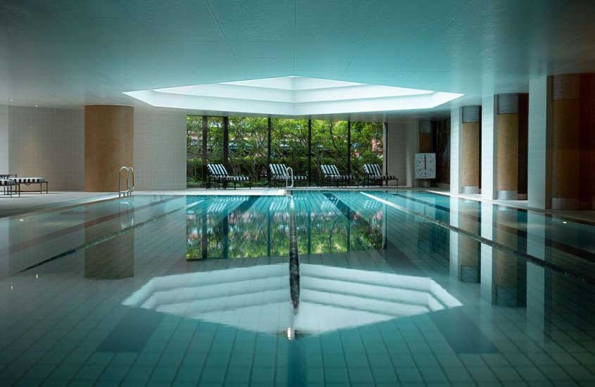 Durante o dia, piscina interna, rodeada por espreguiçadeiras acolchoadas. Ao fundo, janela com vista para uma área verde