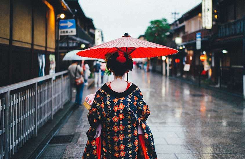 Durante um dia chuvoso, mulher com guarda-chuva vermelho e quimono, caminhando em rua repleta de lojas