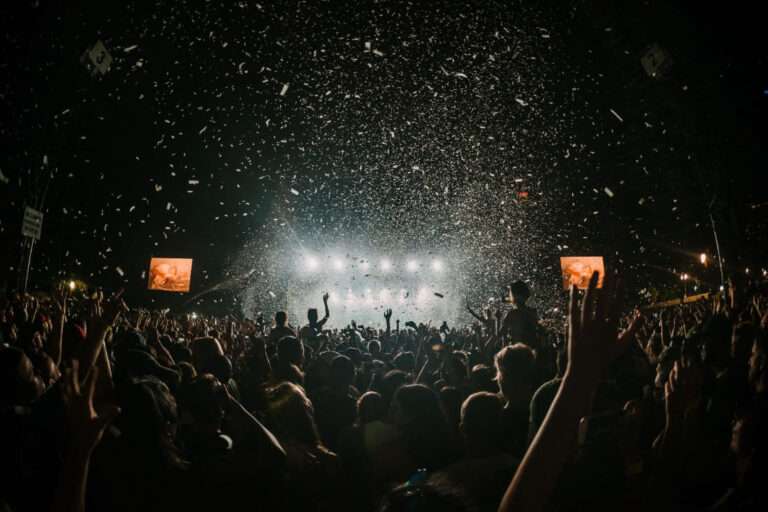 Durante a noite, chuva de papéis coloridos sobre o público que assiste a um show de música em um festival de música. Ao fundo, dois telões ampliam a visão do show