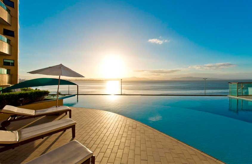 Em final de tarde de sol, espreguiçadeiras, guarda-sol e plantas em frente a piscina com vista para o mar em hotel para passar o réveillon florianópolis 2024