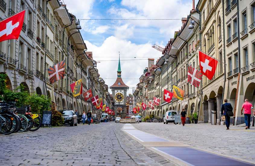 Durante o dia, vários prédios com bandeiras da suíça e da cidade, motos bicicletas, pessoas e carros em uma rua de Altstadt, um lugar onde ficar em Berna