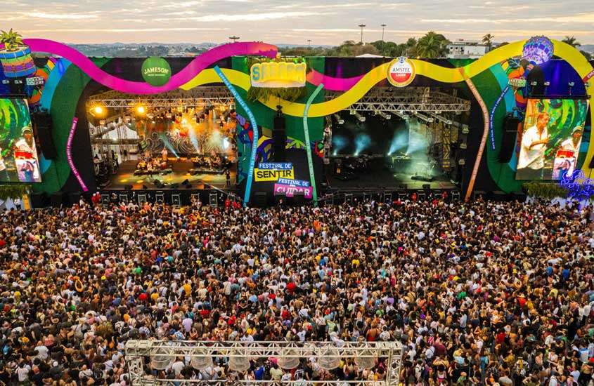 Durante o dia, vista aérea de multidão assistindo shows em festival de musica com dois palcos coloridos