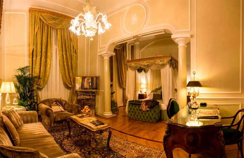 Quarto de hotel em Bolonha com cama com dossel, luminária elegante, janela grande acortinada, sofá, poltrona, mesa de centro, área de trabalho e tapete