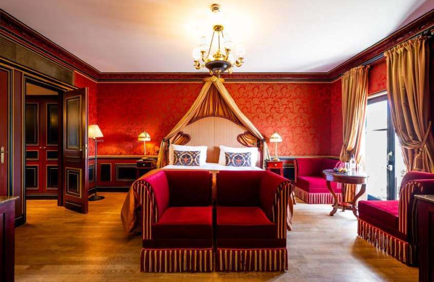 Quarto de hotel em Bordeaux com cama de casal, sofá, divã, cadeiras, mesa, luminárias, lustre ornamentado e janela grande acortinada