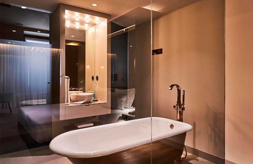 Banheiro de hotel, com banheira, pia moderna com e box refletindo cama de casal de suíte de hotel no porto