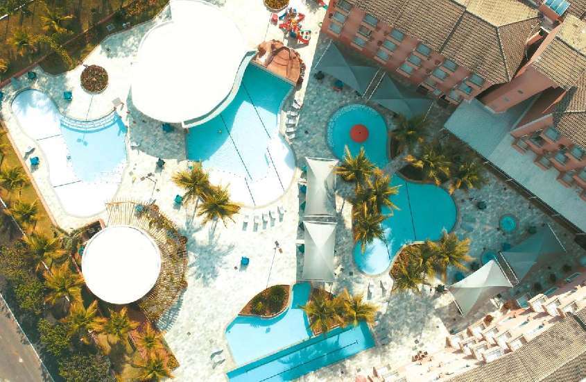 Em dia de sol, vista aérea da área de lazer de hotel com piscinas, árvores ao redor, espreguiçadeiras e playground