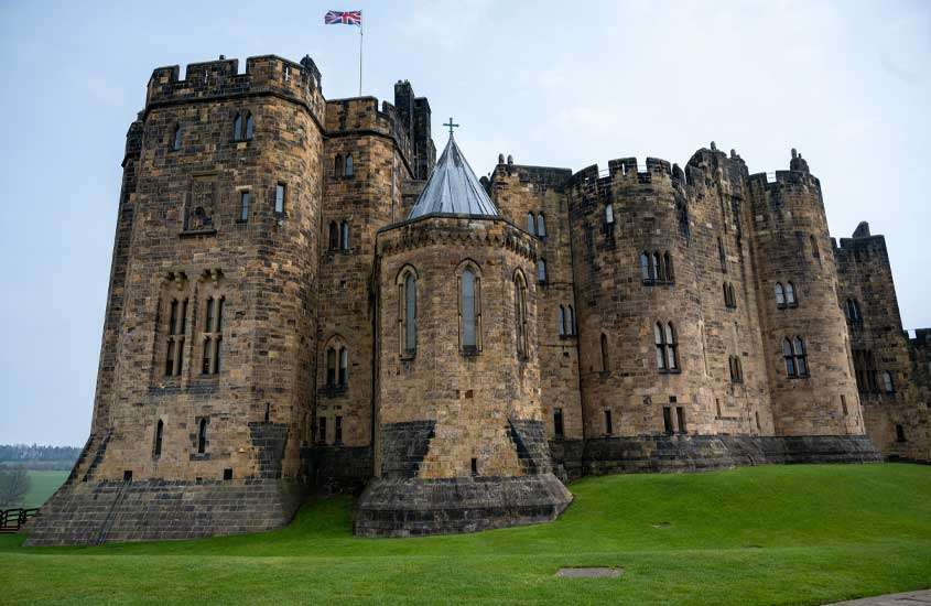 Em dia nublado, castelo britânico com área gramada na frente que foi usado com cenário de filme Harry Potter