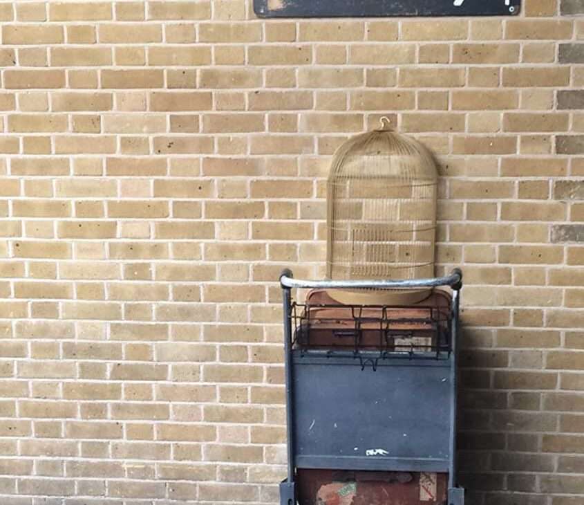Parede de tijolos com placa de Plataforma 9 3/4 e carrinho de malas com gaiola usado como cenário de filme Harry Potter