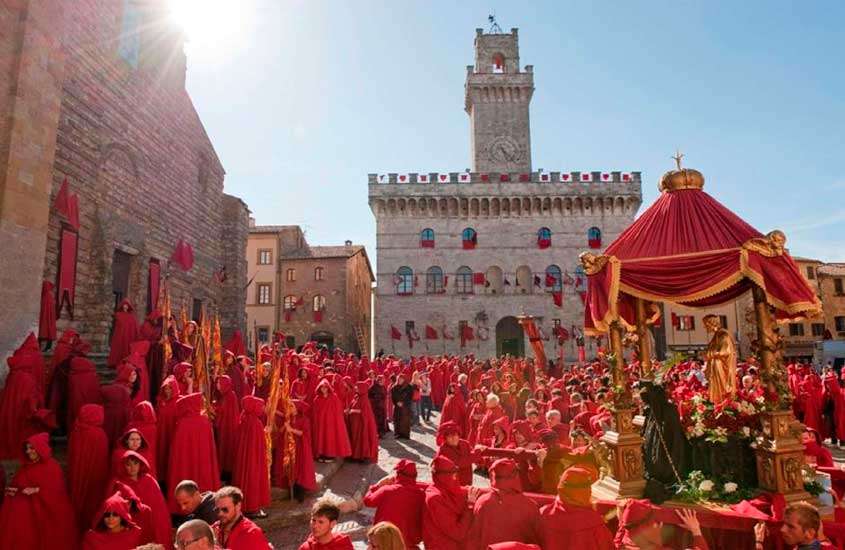Em dia de sol, Centro histórico de Montepulciano, que foi usado como cenário de filme da saga Crepúsculo, com pessoas vestidas em túnicas vermelhas e construções de pedras atrás