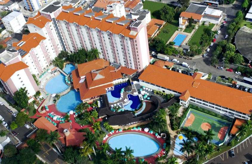 Em dia de sol, vista aérea de hotel para lua de mel em caldas novas com piscinas, quadra, prédios em volta, árvores e rua ao lado com carros