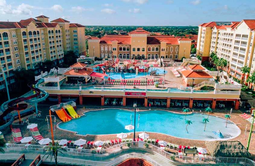 Durante dia de sol, vista aérea de área de lazer de hotele m Orlando com piscina com escorregadores coloridos, guarda-sóis, mesas, cadeiras, espreguiçadeiras, tobogãs e quiosques