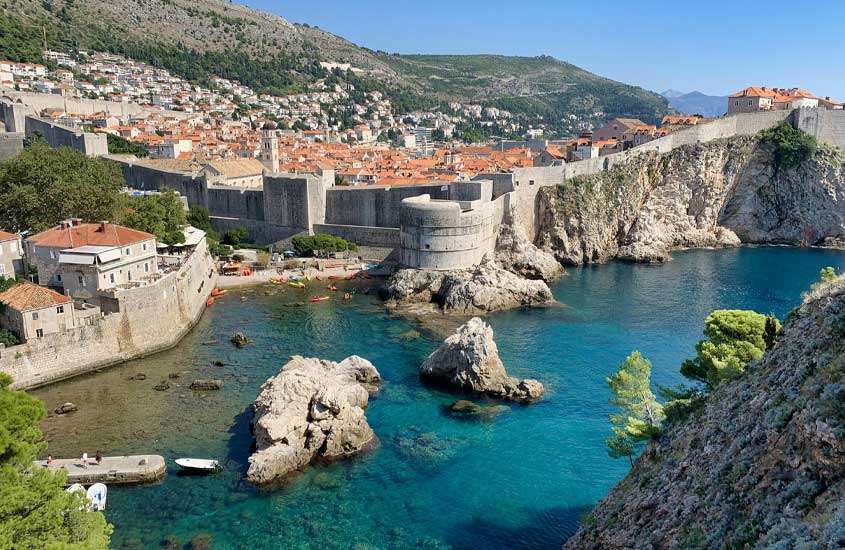 Em dia de sol, vista panorâmica do cenário de série Game of Thrones na Croácia com cidade, floresta atrás e praia com banhistas, paredões de pedra e barcos